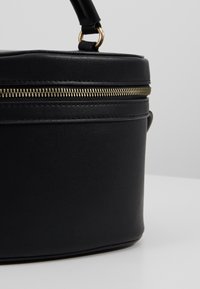 Sac cylindrique en cuir noir avec fermeture éclair dorée et poignée courte attachée par un anneau doré sur fond uni.