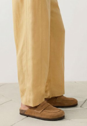 Voeten met bruine suède instap loafers gecombineerd met losse beige broek, staand op een betegelde vloer.