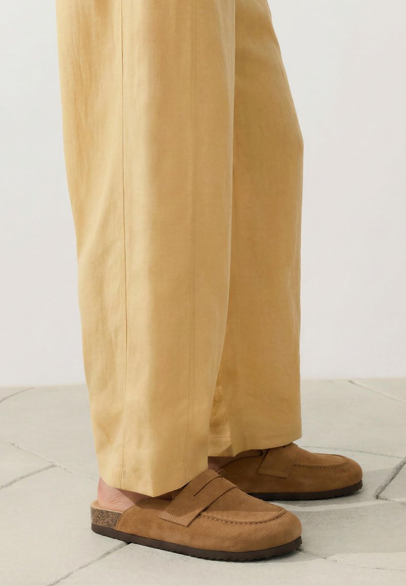 Füße, die braune Wildleder-Slipper tragen, kombiniert mit lockeren beigen Hosen, stehen auf gefliestem Boden.