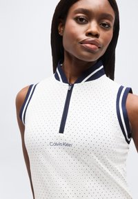 Γυναίκα που φοράει λευκό αμάνικο τοπ Calvin Klein με ναυτικό μπλε επένδυση και μικρές διάτρητες τελείες, αντιμετωπίζοντας προς τα εμπρός με ουδέτερη έκφραση.