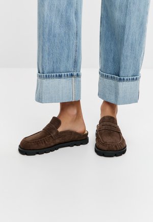 Persona che indossa jeans azzurri risvoltati e mocassini scivolati in pelle scamosciata marrone con suole in gomma nera su un pavimento bianco.