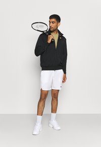 Chaqueta negra con cremallera y cuello, camiseta verde oliva, pantalones cortos blancos y zapatillas blancas. Sosteniendo una raqueta de tenis negra.