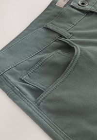 Gros plan d'une poche de pantalon vert avec des coutures détaillées, une passante de ceinture et un bouton métallique sur la surface du tissu.