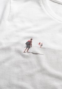 Witte katoenen T-shirt met een print van drie kinderen die over een strand lopen, met lichte schaduw en zachte texturen. Geen aanvullende accenten zichtbaar.