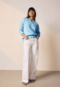 Hele sinine tekstuuriga kampsun V-kaelusega ja soonikkoes äärega, kombinatsioonis valgete laiade pükstega ning mustriliste teravate kontsadega sandaalidega.