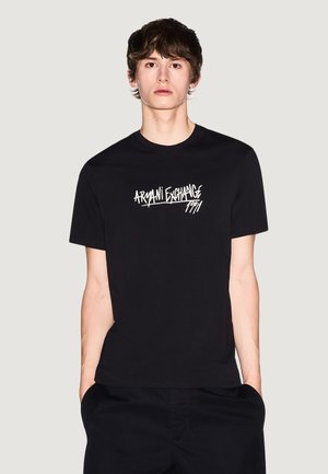 Młody mężczyzna z krótkimi ciemnymi włosami, ubrany w czarną koszulkę Armani Exchange z białym logo, stoi z rękami w kieszeniach.