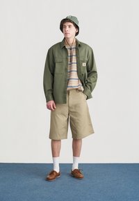 Πράσινο πουκάμισο με μακριά μανίκια, ριγέ μπλουζάκι από κάτω, μπεζ σορτς, καφέ παπούτσια και πράσινο καπέλο τύπου κάδος. Λευκές κάλτσες ολοκληρώνουν την εμφάνιση.