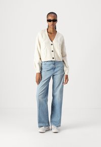 Cardigã de malha canelada em creme com cinco botões, combinado com jeans de perna larga azul claro e ténis brancos. Modelo a usar óculos de sol pretos.