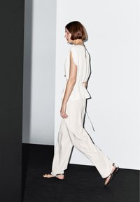 Hvid, løs bluse med korte ærmer og bindebånd bagpå, parret med lysebeige, vide bukser. Sorte flade sandaler fuldender outfittet.