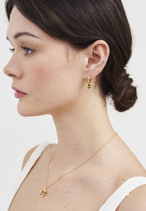 Sieviete ar tumšiem matiem sasietiem bizē, valkā zelta auskarus ar vaska bišu formu un atbilstošu bišu kulonu uz plānas ķēdītes.