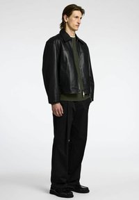 Chaqueta de cuero negra sobre un suéter de punto verde, combinada con pantalones y zapatos negros. La chaqueta tiene cuello con solapa y bolsillos laterales.