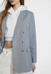 Blå och vit rutmönstrad blazer med dubbelknäppt front, silverfärgade knappar, slagkrage och en enkel bröstficka. Bärs över en vit hoodie.