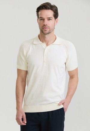 Mężczyzna o krótkich brązowych włosach, ubrany w kremową koszulkę polo z krótkim rękawem i czarne spodnie, stoi na jasnym jednolitym tle.