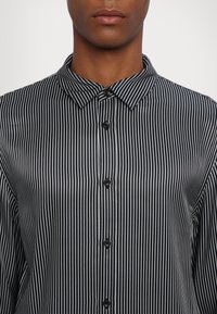 Homme portant une chemise noire à boutons avec de fines rayures blanches verticales et un col pointu, cadré sous le nez.