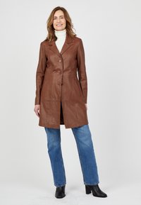 Bruine leren jas met een taillerend ontwerp, enkelknoopsluiting, klep met kraag en zijzakken. Gepaard met blauwe jeans en zwarte laarzen.