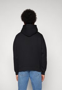 Black hooded sweatshirt with a relaxed fit, featuring a soft texture, ribbed hem, and dropped shoulders. Worn with light blue denim jeans.