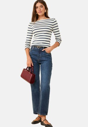 Žena oblečená v bielej a námorníckej modrej pruhovanej mikine s dlhým rukávom, modrých džínsoch, čiernych balerínach, drží malú bordovú kabelku.