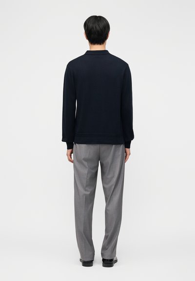 Lühikeste mustade juustega inimene, kes kannab musta pika varrukaga särki ja halli pükse, seisab heleda halli ühtlase taustaga seljaga vaataja poole.