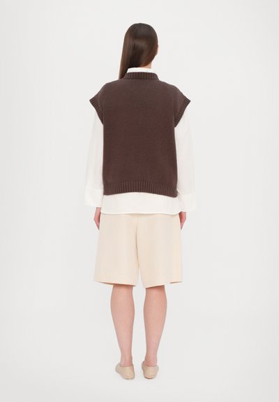 Brauner ärmelloser Pullover mit gerippten Kanten, getragen über einem weißen Hemd mit Knöpfen, kombiniert mit beigen Shorts und neutralen Slipschuhen.