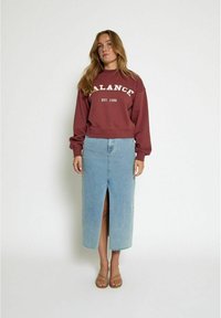 Sudadera burdeos con el texto "BALANCE EST. 1998", combinada con una falda de mezclilla azul claro con una abertura frontal y dobladillo deshilachado. Sandalias beige.
