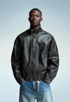 Young man with short hair wearing a black leather jacket over blue jeans with keys hanging from the waistband, posing against a plain background.