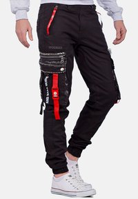 Schwarze Cargo-Hosen mit mehreren Reißverschlusstaschen, Nietenakzenten und roten Gurtdetails. Hergestellt aus strapazierfähigem Stoff mit schmal zulaufenden Bündchen.