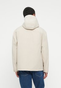 Hombre de espaldas vestido con una chaqueta con capucha beige claro y jeans azules frente a un fondo blanco liso.