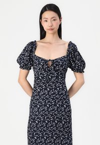Vestido floral azul marinho com mangas bufantes, decote quadrado e um detalhe em forma de olho na parte do busto. Apresenta um padrão de flores brancas por todo o vestido.