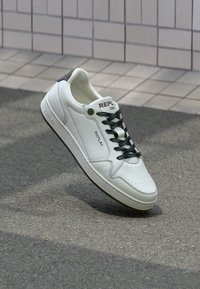 Weiße Leder-Sneaker mit durchlöchtem Zehenbereich, schwarzen Schnürsenkeln und dezentem Branding. Glatte Oberfläche mit niedrigem Profil und Gummisohle.