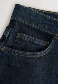 Jeans de denim albastru închis, cu un buzunar frontal cu cusături galbene contrastante și un detaliu din metal pe buzunar.