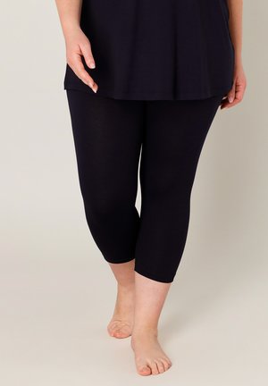 Person trägt schwarze, knielange Leggings und ein schwarzes Oberteil, steht barfuß vor einem einfarbigen hellen Hintergrund.