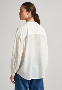 Blusa bianca, leggera, a maniche lunghe con una vestibilità rilassata, maniche arricciate e dettaglio della spalla sul retro. Tessuto morbido e leggermente testurizzato.