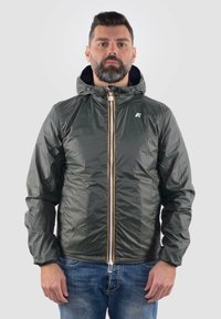Giacca verde, leggera, con finitura lucida, chiusura con zip e cappuccio. Presenta polsini elasticizzati e un logo sul petto. Design semplice.