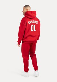 Conjunto de sudadera con capucha roja y pantalones de chándal de tela suave; presenta "SMILODOX 01" en blanco en la parte posterior; corte relajado y capucha ajustable.
