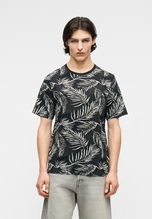 Νεαρός άνδρας με μεσαίου μήκους σκούρα μαλλιά, που φοράει μαύρο μπλουζάκι με λευκά σχέδια από φύλλα φοίνικα και ανοιχτόχρωμα γκρι τζιν, στέκεται μπροστά σε απλό φόντο.