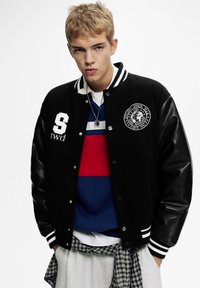 Sort varsityjakke med læderærmer, hvide striber på kraven og manchetterne, prydet med et emblem og bogstavet "S." Lagdelt med stribet top og ternet skjorte.