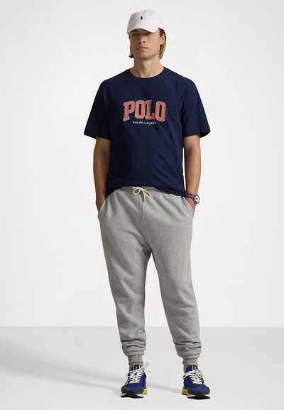 Jūras spēku īsām piedurknēm T-krekls ar sarkano "POLO" logo, kombinēts ar pelēkajiem treniņbiksēm un ziliem sporta apaviem ar dzelteniem akcentiem, kā arī baltu cepuri.