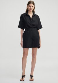 Combinaison courte en lin noir avec col, manches courtes, devant à boutons, taille élastique et ourlets roulés. Portée avec des sandales noires.