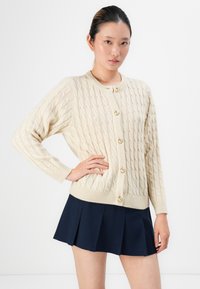 Ung kvinne ikledd en kremfarget cable-knit cardigan med gullknapper og marineblå plisserte skjørt, poserer med hånden på hoften mot en hvit bakgrunn.