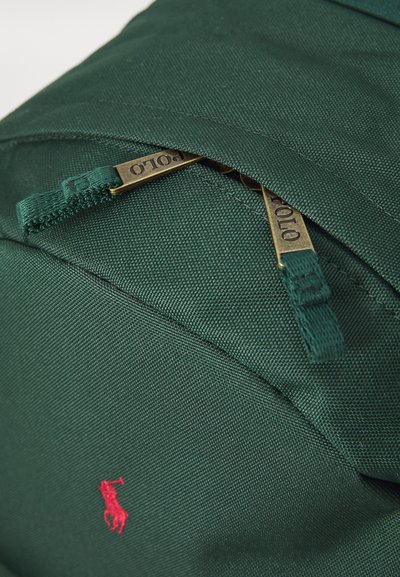Mochila de tela verde con una superficie texturizada, que presenta cremalleras de latón y un logo bordado en rojo. Cremalleras situadas para un acceso fácil.