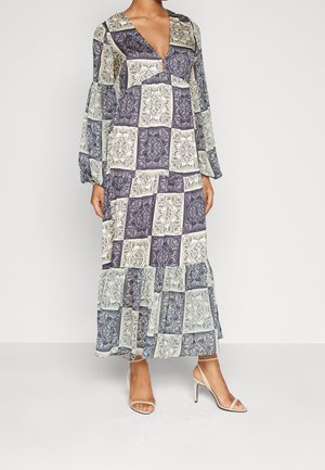 Femme portant une longue robe patchwork ample avec des motifs bleus et crème, encolure en V, manches longues et sandales à talons hauts beiges.