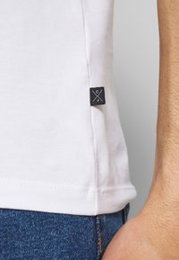 White cotton t-shirt featuring a black label with a logo at the side seam. The fabric is smooth and the cut is fitted. Jeans visible.