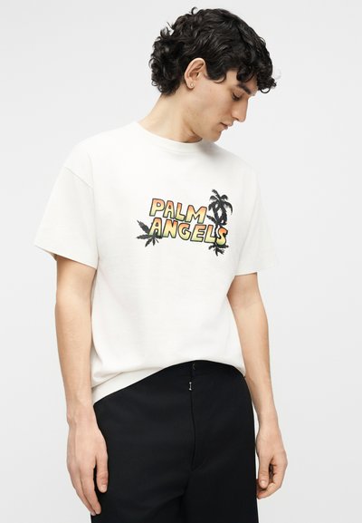 Νεαρός άνδρας με σγουρά σκούρα μαλλιά, φοράει λευκό μπλουζάκι με το λογότυπο "PALM ANGELS" και μαύρο παντελόνι, κοιτάζοντας προς τα κάτω στην αριστερή του πλευρά.