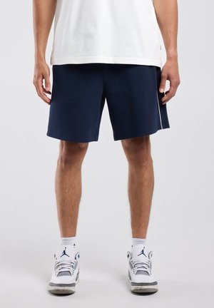 Man draagt marineblauwe korte broek, wit T-shirt, witte sokken en witte sneakers en staat tegen een effen achtergrond.