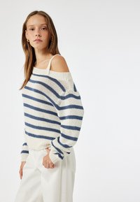 Suéter de rayas crema y azul marino con escote bardot, ajuste suelto, mangas anchas y puños y dobladillo de canalé. Combinado con pantalones blancos.
