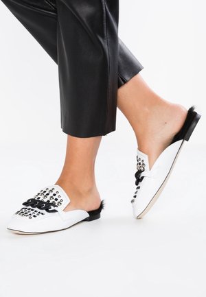 Pieds portant des mocassins blancs sans lacets avec chaîne noire et détails cloutés, associés à un pantalon en cuir noir coupé court.