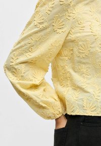 Blouse jaune brodée avec des manches bouffantes et un motif floral texturé. Tissu doux avec des poignets froncés et une coupe courte.