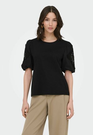 Kobieta ubrana w czarną bluzkę z koronkowymi bufkami na rękawach oraz beżowe spodnie o szerokich nogawkach, stojąca na jednolitym białym tle.