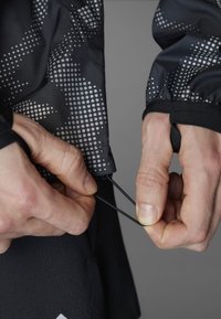 Schwarze Jacke mit reflektierendem Punktmuster, elastischen Bündchen und verstellbarem Kordelzug. Hände passen die Jacke am Saum an.