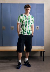 T-shirt blanche avec un motif abstrait vert, col rond, et logo ; shorts cargo noirs ; baskets noires avec accents bleus ; fond de vestiaire.